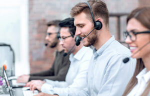 Three male and one female call center workers smiling, sitting in a line with headsets on and typing on their computers.