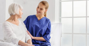 Blonde female nurse assisting a senior female patient in a white room with windows