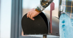 A male wearing a flannel shirt and light blue jeans closes a black mailbox to send a payment