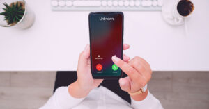 A person in black jeans and a white longsleeve shirt is sitting down at a desk and holding a touchscreen cell phone that has an Unknown phone number calling them