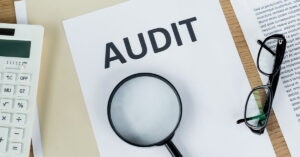 A folder, paper that reads "AUDIT", another piece of paper out of frame that has various text on it, a white calculator, a black pair of reading glasses, and a black magnifying glass, all on top of a wooden table.