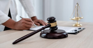 A person is writing with a pen on a clipboard with the scales of justice and a wooden gavel on the table