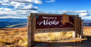 A wooden road sign reads, "Welcome to Alaska" which overlooks a mountainous landscape and a partly cloudy sky