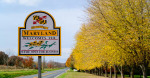 A road sign that says, "Maryland Welcomes You" and the background features a long country road and fall-colored leaves