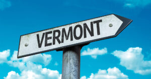 A road sign in the shape of an arrow pointing to the right says "Vermont" with a blue sky with clouds in the background