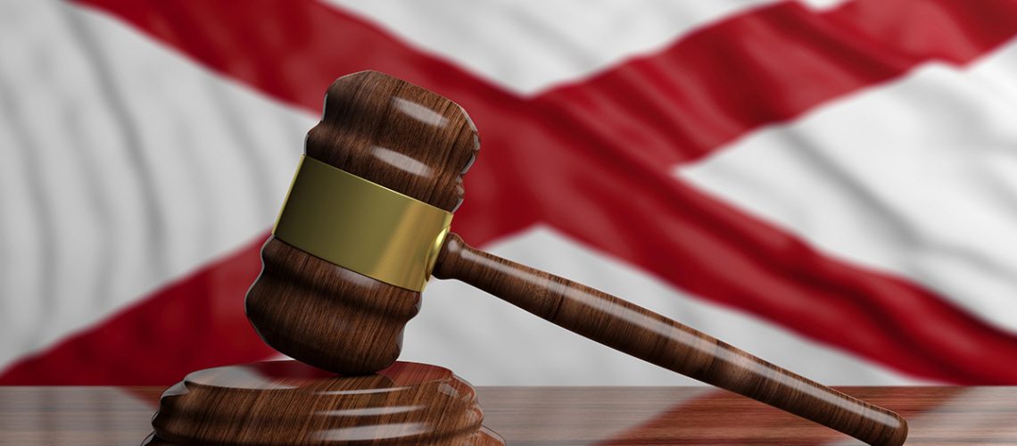 The Alabama flag is hanging in the background of a wooden gavel on top of a wooden table