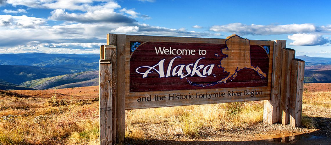 A wooden road sign reads, "Welcome to Alaska" which overlooks a mountainous landscape and a partly cloudy sky