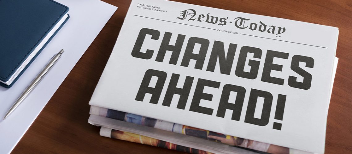 "Changes Ahead!" printed on a newspaper sitting on a wooden desk next to a blue note pad and silver pen