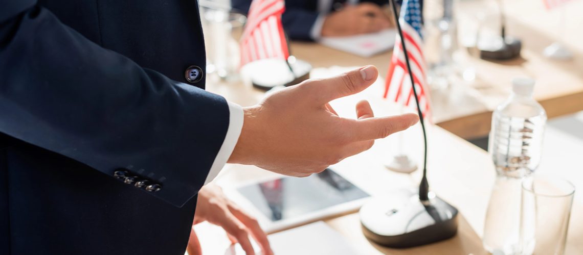 A man in a suit is gesturing with his hands and a table with notebooks, water bottles, microphones, and mini American flags on top