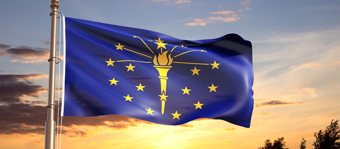 The blue and yellow Indiana flag waves on a flagpole in front of a sunset with clouds and tree silhouettes.