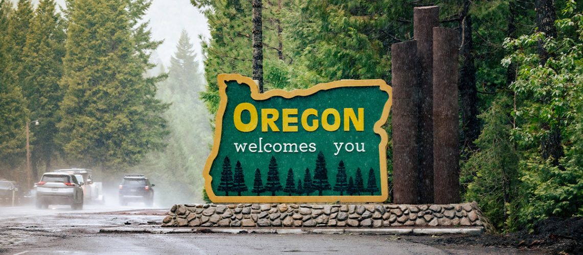 The state of Oregon road sign welcomes drivers on a rainy day with tall green pine trees in the background
