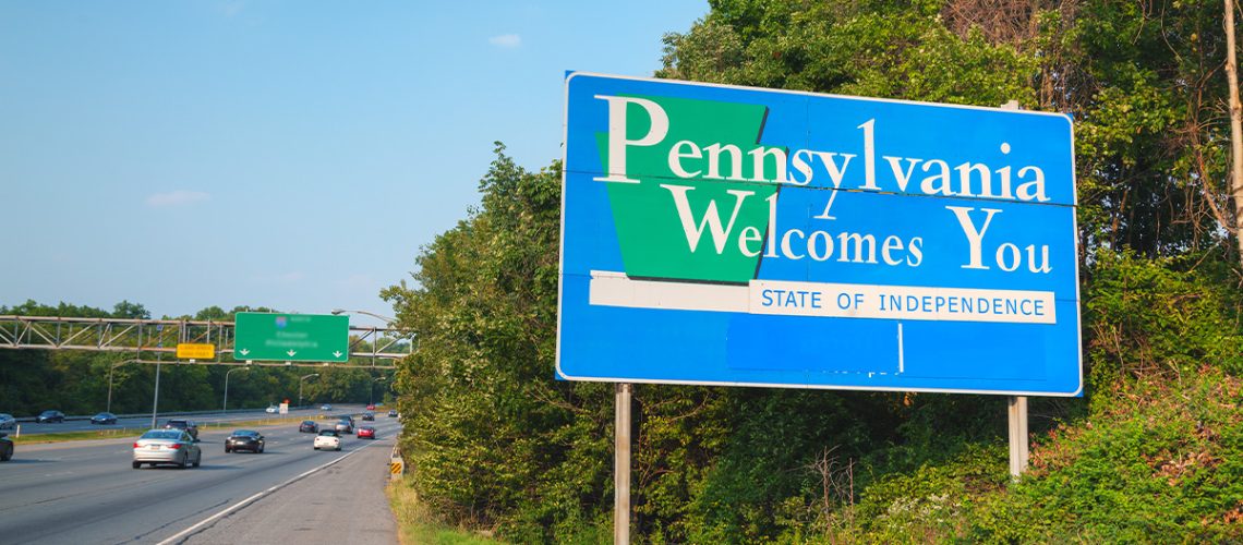 A Pennsylvania road sign says "Pennsylvania Welcomes You" with the highway and several cars in the background