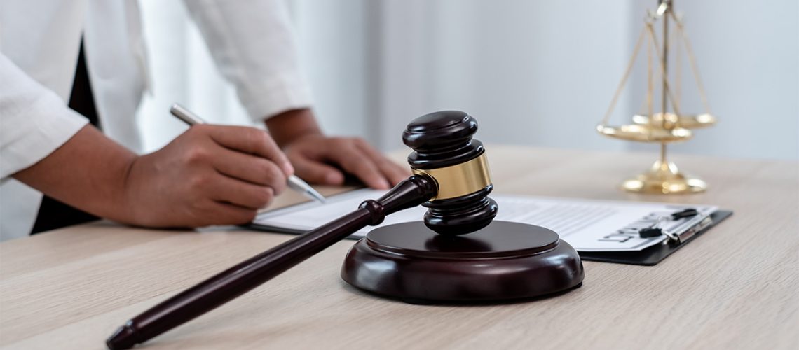 A person is writing with a pen on a clipboard with the scales of justice and a wooden gavel on the table