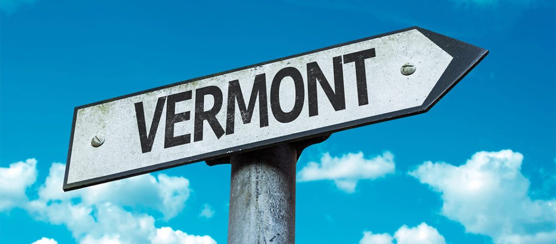 A road sign in the shape of an arrow pointing to the right says "Vermont" with a blue sky with clouds in the background